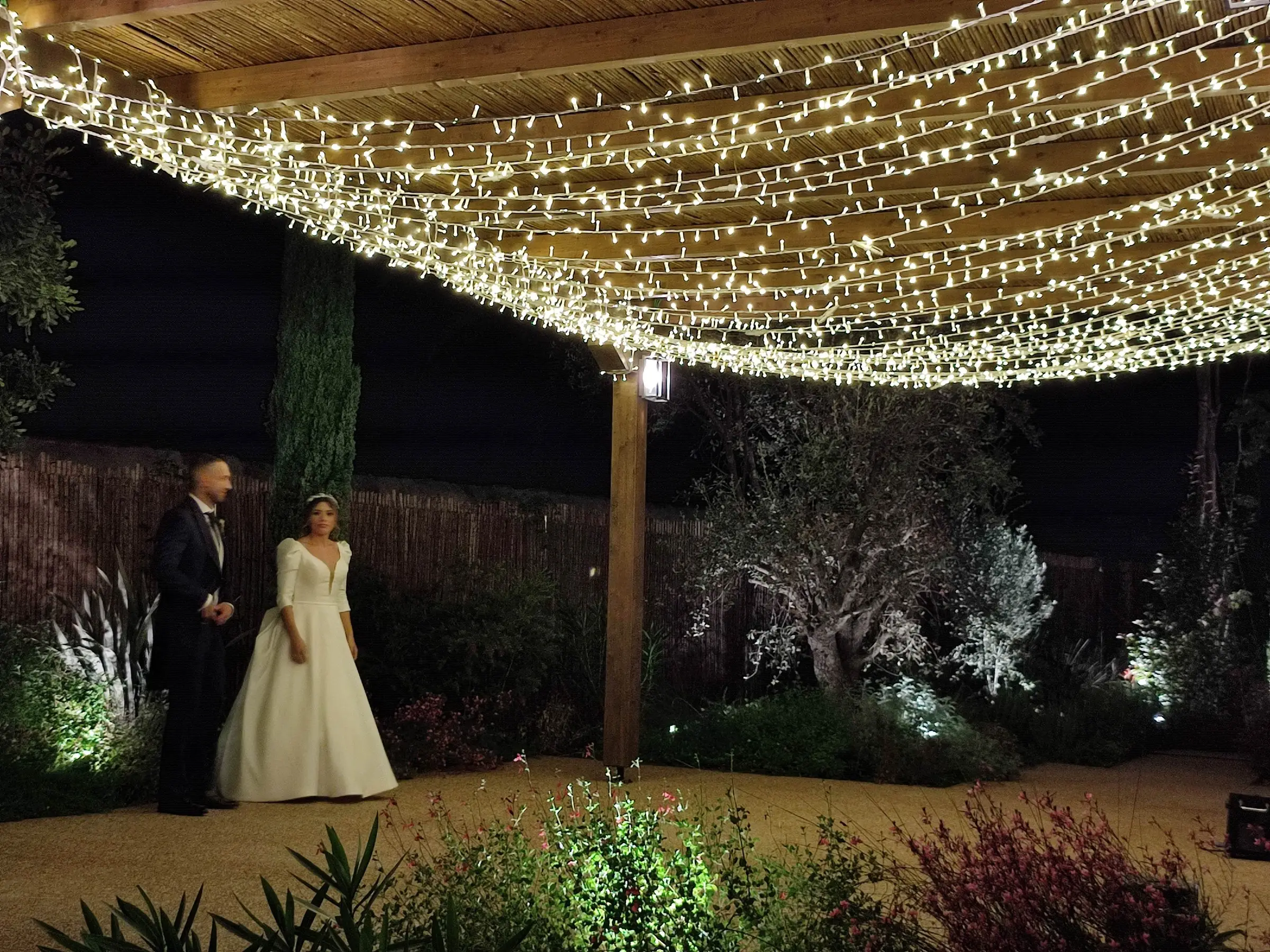 De noche, los novios pasean bajo un techo de guirnaldas de luces sobre una pérgola de madera, entre olivos y lavanda iluminados
