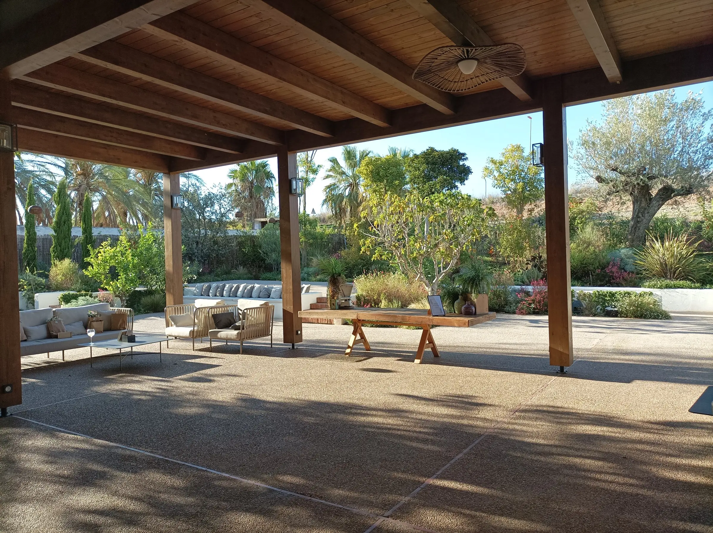 Un porche de vigas de madera abierto a un jardín con olivos, palmeras y flores, con sofás y una mesa larga