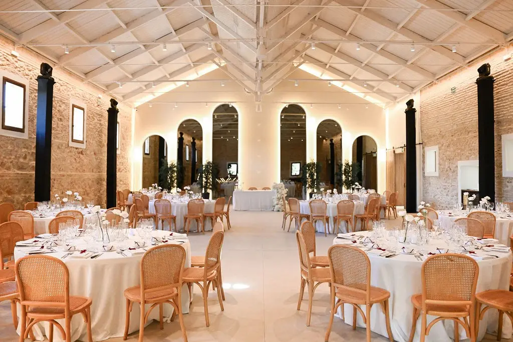 Mesas redondas con mantel blanco y sillas de rejilla en el salón, con columnas negras y ventanales de arco al fondo